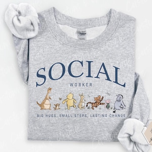 Pu&ograve; includere: Felpa grigio chiaro con la scritta blu navy "SOCIAL WORKER" ad arco nella parte superiore. Sotto il testo c'&egrave; una fila di personaggi dei cartoni animati. La parte inferiore della maglietta recita "BIG HUGS, SMALL STEPS, LASTING CHANGE."