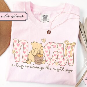 May include: A light pink t-shirt with the word "NAGH" in a pink heart pattern, featuring a cartoon bear and a honey pot. The text "a hug is always the right size" is below the word. A gold necklace and a brown hat are also visible.