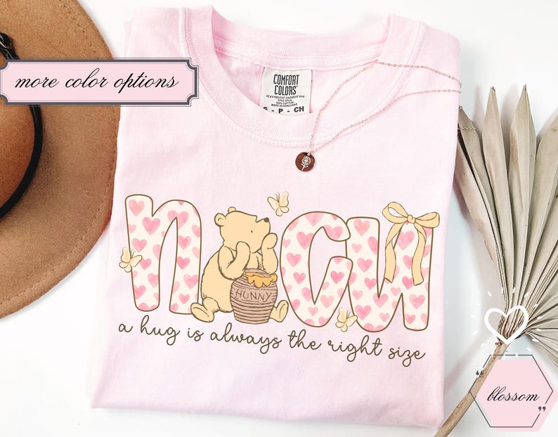 May include: A light pink t-shirt with the word "NAGH" in a pink heart pattern, featuring a cartoon bear and a honey pot. The text "a hug is always the right size" is below the word. A gold necklace and a brown hat are also visible.