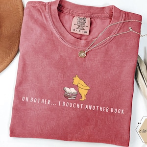 May include: A red Comfort Colors t-shirt featuring a graphic of Winnie the Pooh and books, with the text "OH BOTHER... I BOUGHT ANOTHER BOOK." A silver necklace with a pendant is draped across the shirt. A brown hat and decorative leaves are in the background.