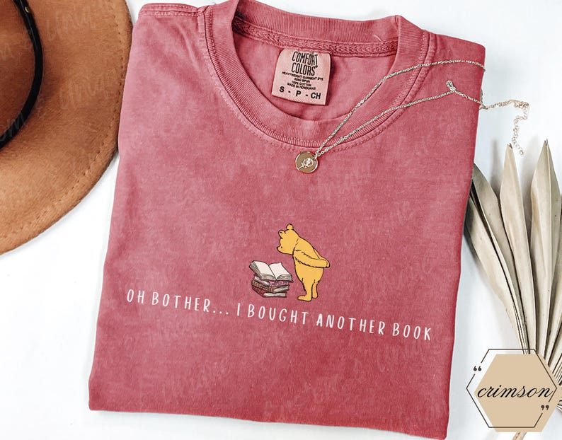 May include: A red Comfort Colors t-shirt featuring a graphic of Winnie the Pooh and books, with the text "OH BOTHER... I BOUGHT ANOTHER BOOK." A silver necklace with a pendant is draped across the shirt. A brown hat and decorative leaves are in the background.