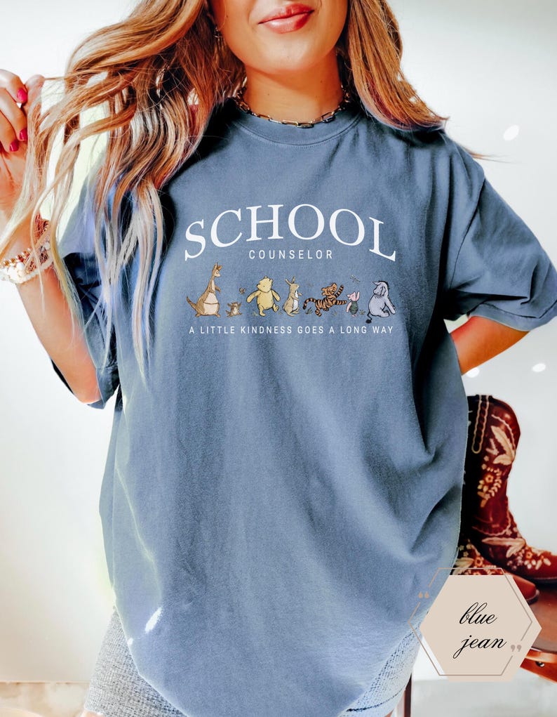 May include: A blue-gray crewneck t-shirt featuring the white text SCHOOL COUNSELOR above a graphic of Winnie the Pooh characters and the text A LITTLE KINDNESS GOES A LONG WAY. The shirt has short sleeves and a relaxed fit with a vintage-style wash.