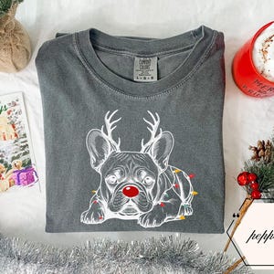 May include: A charcoal gray t-shirt featuring a white illustration of a French Bulldog with reindeer antlers and a red nose. The dog is adorned with Christmas lights. A red mug with "Merry & Bright" is visible.
