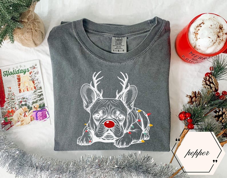 May include: A charcoal gray t-shirt featuring a white illustration of a French Bulldog with reindeer antlers and a red nose. The dog is adorned with Christmas lights. A red mug with "Merry & Bright" is visible.