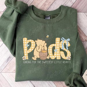 Puede incluir: Una sudadera verde militar con la palabra "Pods" en letras de cuadros amarillos, con un oso de dibujos animados y un tarro de miel. El texto "Caring for the sweetest little hearts" está debajo. La sudadera tiene mangas largas.