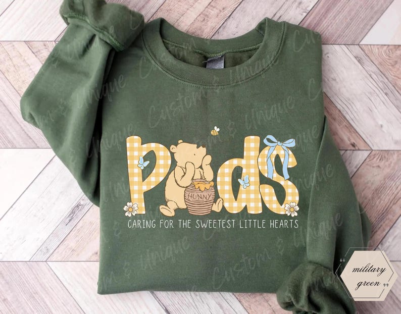 May include: A military green sweatshirt with the word "Pods" in yellow plaid letters, featuring a cartoon bear and honey pot. The text "Caring for the sweetest little hearts" is below the graphic. The sweatshirt has long sleeves.