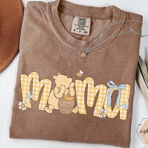 May include: Brown t-shirt with the word "mama" in yellow gingham print, featuring Winnie the Pooh, a honey pot, and a bee. A silver necklace with a small pendant is on the shirt. The shirt has a Comfort Colors tag.