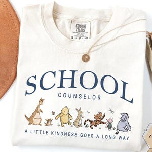May include: An ivory t-shirt featuring the dark blue text SCHOOL COUNSELOR and a graphic of Winnie the Pooh characters. Below the characters, the text reads A LITTLE KINDNESS GOES A LONG WAY. The shirt has a Comfort Colors label in the neck area.