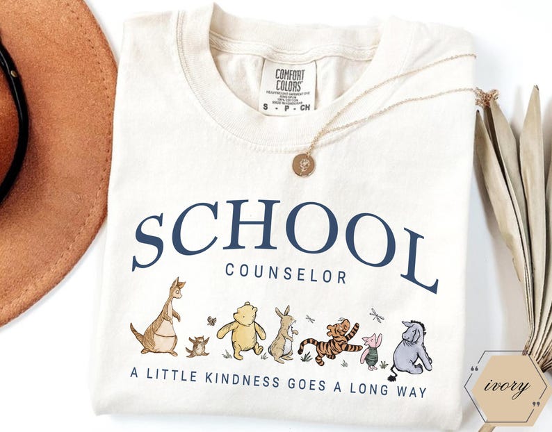 May include: An ivory t-shirt featuring the dark blue text SCHOOL COUNSELOR and a graphic of Winnie the Pooh characters. Below the characters, the text reads A LITTLE KINDNESS GOES A LONG WAY. The shirt has a Comfort Colors label in the neck area.
