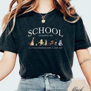 May include: A black short-sleeve t-shirt featuring the white text SCHOOL COUNSELOR above a graphic of Winnie the Pooh characters and the text A LITTLE KINDNESS GOES A LONG WAY. The shirt has a crew neckline and a relaxed fit with a vintage-style wash.