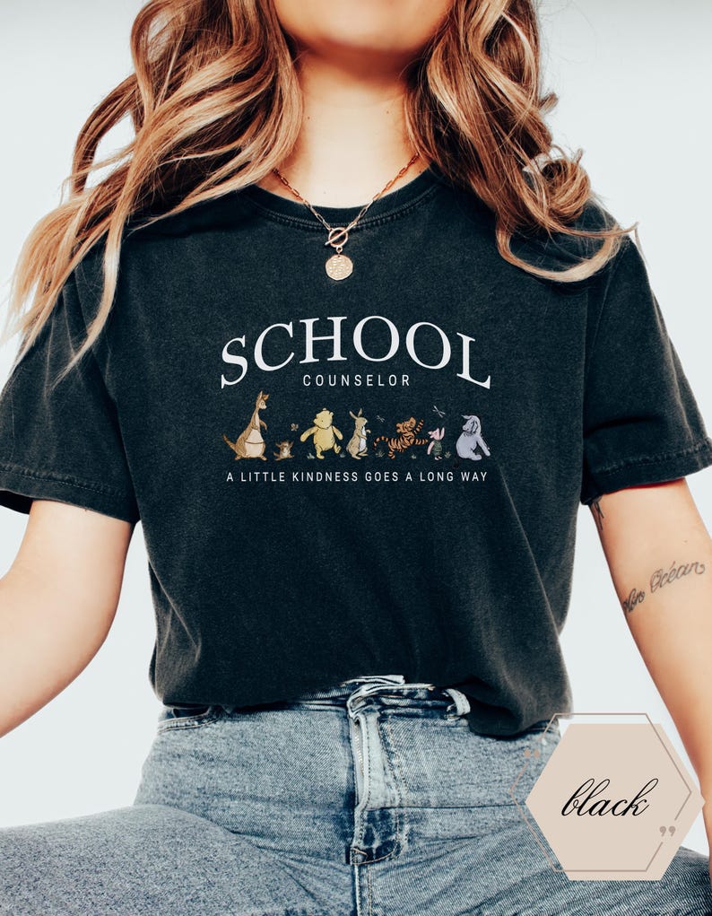 May include: A black short-sleeve t-shirt featuring the white text SCHOOL COUNSELOR above a graphic of Winnie the Pooh characters and the text A LITTLE KINDNESS GOES A LONG WAY. The shirt has a crew neckline and a relaxed fit with a vintage-style wash.