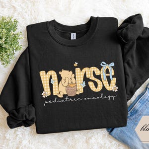 Peut inclure: Sweat-shirt noir avec un motif graphique. Le motif pr&eacute;sente le mot "nurse" en imprim&eacute; vichy jaune, un ours de dessin anim&eacute; et les mots "pediatric oncology". Un n&oelig;ud bleu et des fleurs sont &eacute;galement inclus dans le motif.