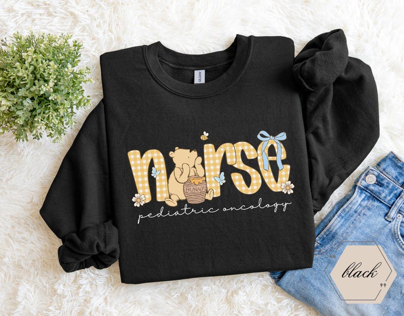 Peut inclure: Sweat-shirt noir avec un motif graphique. Le motif pr&eacute;sente le mot "nurse" en imprim&eacute; vichy jaune, un ours de dessin anim&eacute; et les mots "pediatric oncology". Un n&oelig;ud bleu et des fleurs sont &eacute;galement inclus dans le motif.