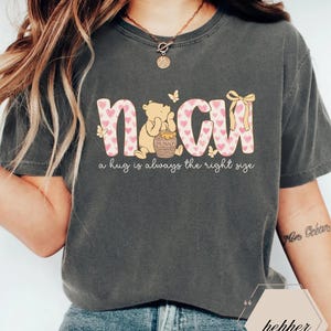 May include: Dark gray t-shirt with the word "NCW" in pink letters with heart patterns. A cartoon bear is sitting next to a honey pot. The text "a hug is always the right size" is below the letters. Butterflies and a bow are also featured.