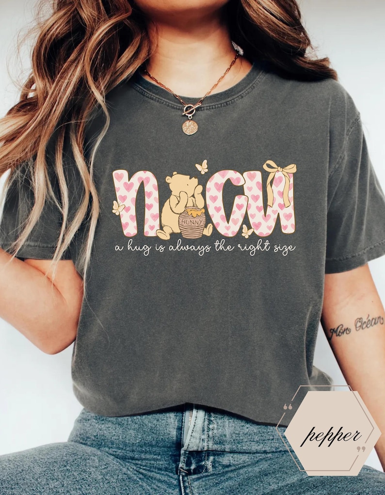 May include: Dark gray t-shirt with the word "NCW" in pink letters with heart patterns. A cartoon bear is sitting next to a honey pot. The text "a hug is always the right size" is below the letters. Butterflies and a bow are also featured.