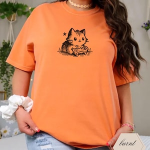 May include: Burnt orange t-shirt featuring a black graphic of a cat playing a video game. The cat is holding a game controller. The shirt has short sleeves and a crew neck. The text "burnt orange" is in a hexagon shape.