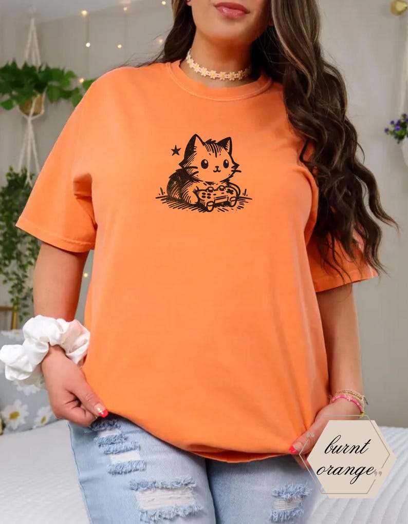 May include: Burnt orange t-shirt featuring a black graphic of a cat playing a video game. The cat is holding a game controller. The shirt has short sleeves and a crew neck. The text "burnt orange" is in a hexagon shape.