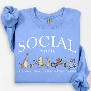 Pu&ograve; includere: Felpa blu Carolina con la scritta bianca "SOCIAL WORKER" e personaggi dei cartoni animati. Sotto, le parole "BIG HUGS, SMALL STEPS, LASTING CHANGE". La felpa ha un girocollo e maniche lunghe.