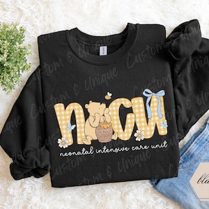 May include: Black sweatshirt with "NICU" in yellow gingham letters, featuring a cartoon bear with a honey pot, a bee, and floral accents. The text "neonatal intensive care unit" is below the letters. A blue bow is on the letter "U".