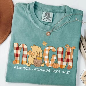 May include: A light green Comfort Colors t-shirt featuring the letters "NICU" in a plaid design with a cartoon bear, pumpkin, and autumn leaves. The text "neonatal intensive care unit" is printed below the letters. A silver necklace is draped across the shirt.