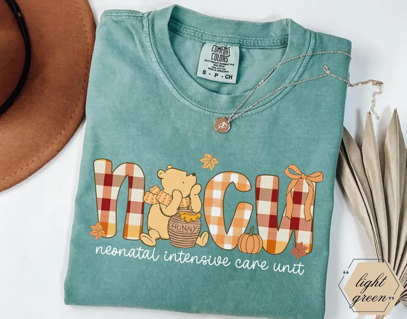 May include: A light green Comfort Colors t-shirt featuring the letters "NICU" in a plaid design with a cartoon bear, pumpkin, and autumn leaves. The text "neonatal intensive care unit" is printed below the letters. A silver necklace is draped across the shirt.