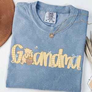 Pode incluir: Uma camiseta Comfort Colors azul claro com a palavra "Vovó" em letras xadrez amarelas. O design inclui um urso de desenho animado, um pote de mel, flores e borboletas. Um colar de ouro com um pingente está sobre a camiseta.