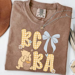 May include: Brown Comfort Colors t-shirt with a Winnie the Pooh graphic and the text "BC BA" in yellow gingham letters. The design includes a blue bow, butterflies, and flowers. A silver necklace and a brown hat are also visible.