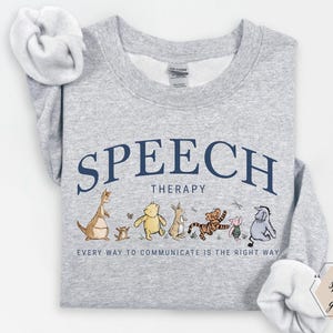 May include: A light grey sweatshirt featuring the words "SPEECH THERAPY" in navy blue arched text. Below, a line of cartoon characters. The text "EVERY WAY TO COMMUNICATE IS THE RIGHT WAY" is below the characters.