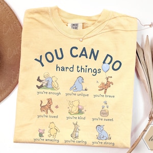 May include: A butter yellow t-shirt featuring the text YOU CAN DO hard things and illustrations of Winnie the Pooh characters. Below the text are nine small scenes with captions including you're enough, unique, brave, loved, kind, sweet, amazing, caring, and strong.