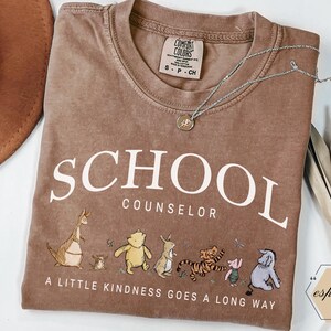 May include: A brown short-sleeved t-shirt with the text SCHOOL COUNSELOR and A LITTLE KINDNESS GOES A LONG WAY printed in white. The shirt features a graphic of Winnie the Pooh characters including Roo, Piglet, Pooh, Rabbit, Tigger, and Eeyore.