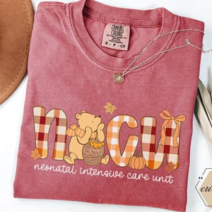 May include: A maroon Comfort Colors t-shirt with a graphic that reads "NICU" in plaid letters, featuring a cartoon bear with a honey pot and a pumpkin. The words "neonatal intensive care unit" are below the graphic.
