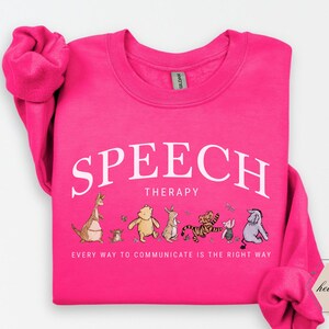 May include: A bright pink sweatshirt with the words "SPEECH THERAPY" in white arched text. Below, the phrase "EVERY WAY TO COMMUNICATE IS THE RIGHT WAY" is printed. A line of cartoon characters are featured below the text.