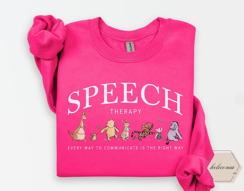 May include: A bright pink sweatshirt with the words "SPEECH THERAPY" in white arched text. Below, the phrase "EVERY WAY TO COMMUNICATE IS THE RIGHT WAY" is printed. A line of cartoon characters are featured below the text.