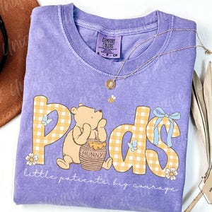 May include: A lavender Comfort Colors t-shirt featuring a Winnie the Pooh design with the word "Pads" in yellow gingham letters. The shirt includes the text "little patients, big courage" and a gold necklace.