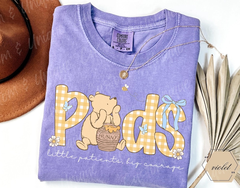 May include: A lavender Comfort Colors t-shirt featuring a Winnie the Pooh design with the word "Pads" in yellow gingham letters. The shirt includes the text "little patients, big courage" and a gold necklace.