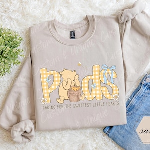 May include: A sand-colored sweatshirt with a graphic featuring the word "Pods" in yellow gingham letters, a cartoon bear, and the text "Caring for the sweetest little hearts." The design includes a bee, flowers, and a blue bow.