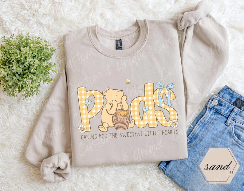 May include: A sand-colored sweatshirt with a graphic featuring the word "Pods" in yellow gingham letters, a cartoon bear, and the text "Caring for the sweetest little hearts." The design includes a bee, flowers, and a blue bow.