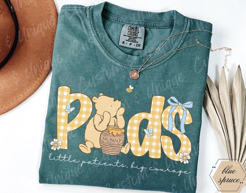 May include: Teal Comfort Colors t-shirt featuring a Winnie the Pooh design with the word "Pods" in yellow gingham letters. The design includes a bear, honey pot, butterflies, and the text "little patients, big courage."