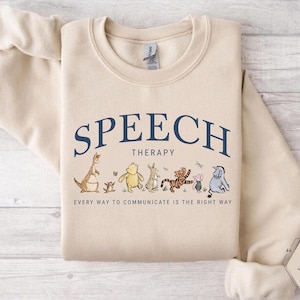 May include: Sand-colored sweatshirt with the words "SPEECH THERAPY" in navy blue arched across the top. Below the text are cartoon characters. The text "EVERY WAY TO COMMUNICATE IS THE RIGHT WAY" is printed below the characters.
