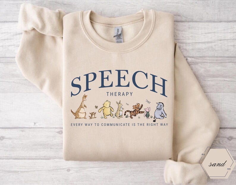 May include: Sand-colored sweatshirt with the words "SPEECH THERAPY" in navy blue arched across the top. Below the text are cartoon characters. The text "EVERY WAY TO COMMUNICATE IS THE RIGHT WAY" is printed below the characters.