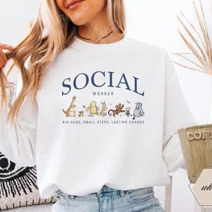 Pu&ograve; includere: Felpa bianca con la scritta "SOCIAL WORKER" in blu navy, con personaggi dei cartoni animati. Sotto il testo c'&egrave; la frase "BIG HUGS, SMALL STEPS, LASTING CHANGE". Una tazza di caff&egrave; &egrave; tenuta in mano.