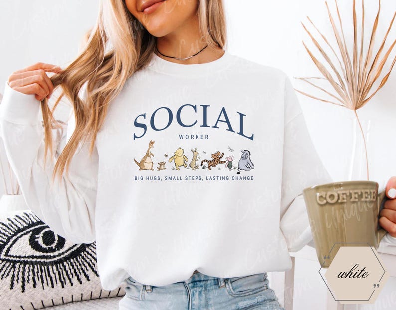 Pu&ograve; includere: Felpa bianca con la scritta "SOCIAL WORKER" in blu navy, con personaggi dei cartoni animati. Sotto il testo c'&egrave; la frase "BIG HUGS, SMALL STEPS, LASTING CHANGE". Una tazza di caff&egrave; &egrave; tenuta in mano.