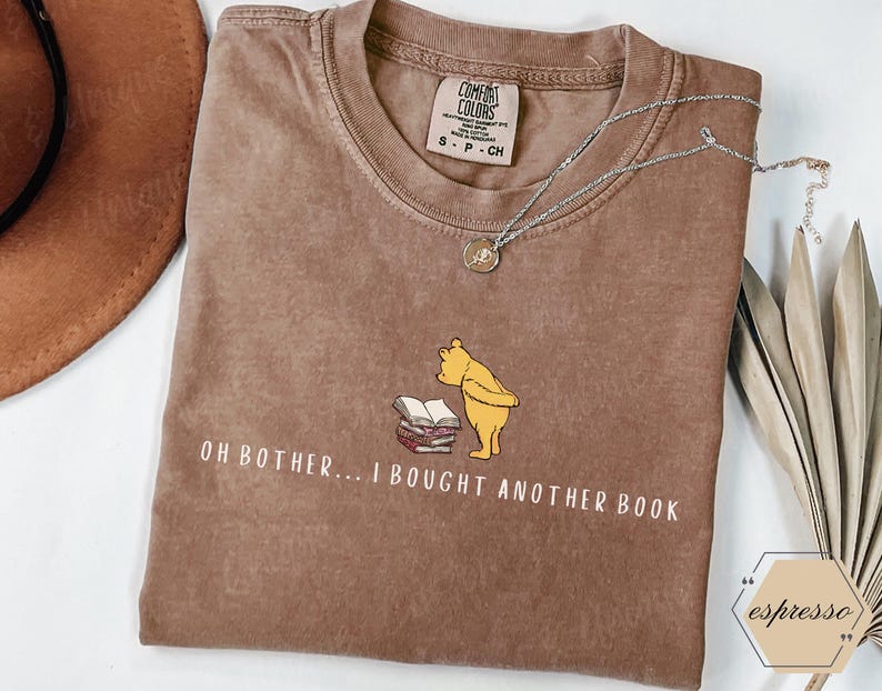 May include: A brown t-shirt with the phrase "OH BOTHER... I BOUGHT ANOTHER BOOK" and a cartoon illustration of Winnie the Pooh. A silver necklace with a pendant is draped across the shirt.