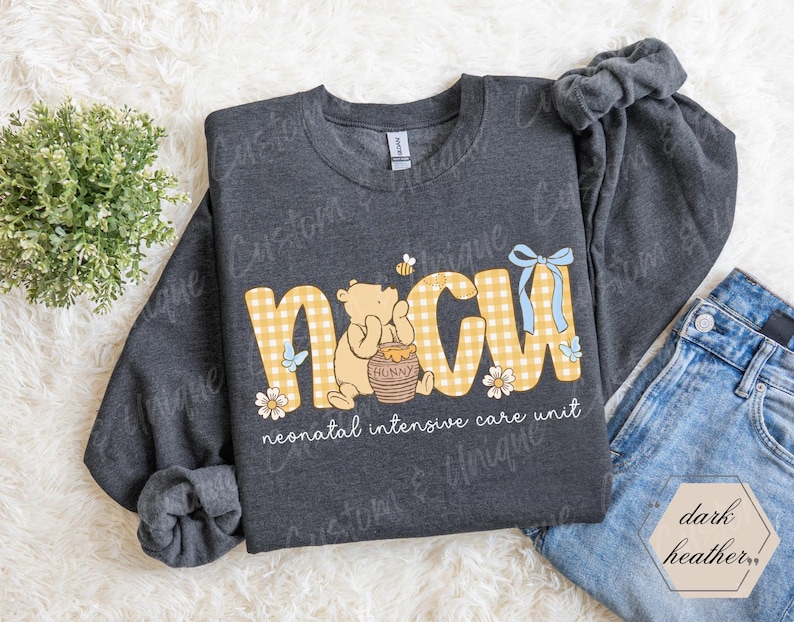 May include: Dark heather gray sweatshirt with the letters "NICU" in yellow gingham print, featuring a cartoon bear, honey pot, and flowers. The words "neonatal intensive care unit" are printed below the letters.
