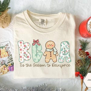 Könnte beinhalten: Ein elfenbeinfarbenes T-Shirt mit einer Grafik, die den Text "BEAR" und "Tis the Season to Reinforce" zeigt. Der Buchstabe B hat ein Weihnachtsmuster, das E ist eine grüne Form mit roter Schleife, das A ein Lebkuchenmann und das R ein Tannenzweigmuster.