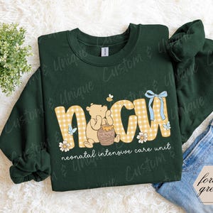 May include: Forest green sweatshirt with "NICU" in yellow gingham letters, Winnie the Pooh graphic, and text "neonatal intensive care unit." Features a blue bow and floral accents. A cozy, long-sleeved top.
