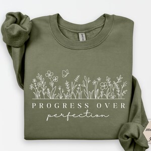 以下が含まれることがあります： 白い花の模様と「PROGRESS OVER perfection」の文字がプリントされたオリーブグリーンのスウェットシャツ。スウェットシャツはクルーネックと長袖です。色は「ミリタリーグリーン」と表示されています。