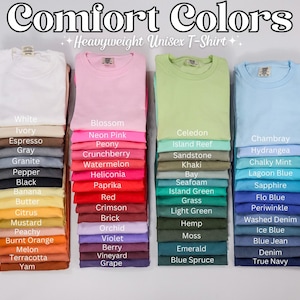 May include: Stacks of folded Comfort Colors heavyweight unisex t-shirts in various colors. The image displays a range of hues, including white, pink, green, and blue, with color names listed below each stack. The text "Comfort Colors" is at the top.