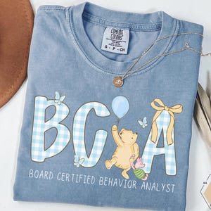 以下が含まれることがあります： 水色のComfort Colors Tシャツ。薄い青色のギンガムチェック模様の「BCA」の文字と、風船を持った漫画のクマが描かれています。「BOARD CERTIFIED BEHAVIOR ANALYST」の文字が下にプリントされています。金のネックレスがシャツの上にかけられています。
