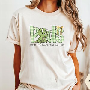 May include: Ivory t-shirt with the word "Peds" in green plaid letters and a cartoon dinosaur. The shirt also features the phrase "Caring for Rawr-some Patients" in black text. A yellow bow accents the "ds" in "Peds".